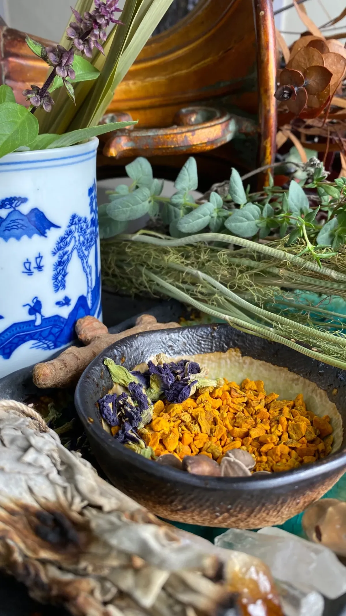 Fresh and dried herbs cover a table. Dried sage, tea leaves, and crystals are in the forefront of the image. A white ceramic mug with blue figures, trees, and mountains holds fresh lemongrass.