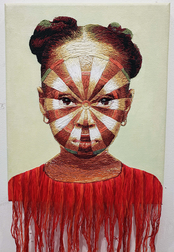 An embroidered portrait on canvas of a young medium-dark skinned girl from the shoulders up. She looks directly at the viewer; her face has the pattern of a dart board on it. Her shirt is made from bright red thread, which hangs off the bottom of the canvas loosely. 