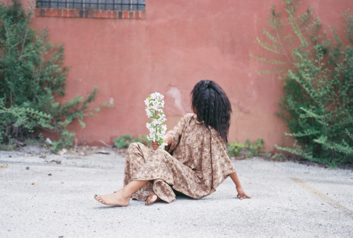 A dark-skinned woman in a flowing brown patterned tunic sits barefoot on a cement ground, leaning back on her left hand. In her right hand, she holds a stalk of white flowers; her head is turned away from the camera and faces a pink clay wall behind her.