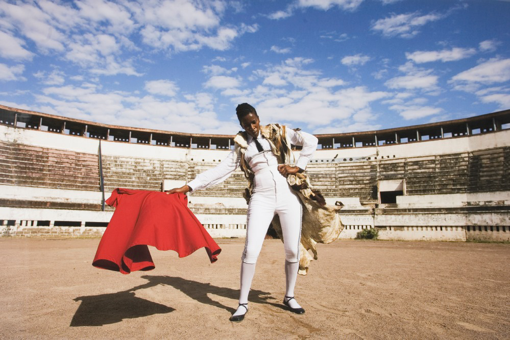 A dark-skinned woman wearing a white, skin-fitting bullfighter’s suit flourishes a red cloth in an empty wooden stadium. A bright blue sky with wispy clouds stretches above her; the photograph is taken in harsh daylight, and she casts a dark shadow behind her.