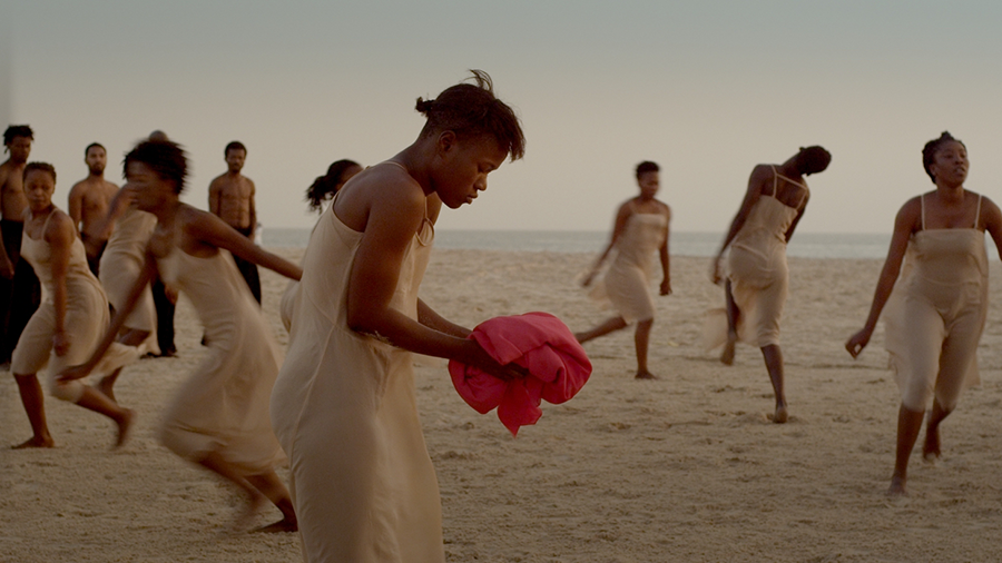 A group of dark-skinned women wearing tan-colored slip dresses run gracefully on an empty beach at twilight. One woman in the center stands still, her shoulders hunched over a pink bolt of cloth in her hands. Behind them, dark-skinned men stand without shirts stand and watch. The colors are muted and dream-like.