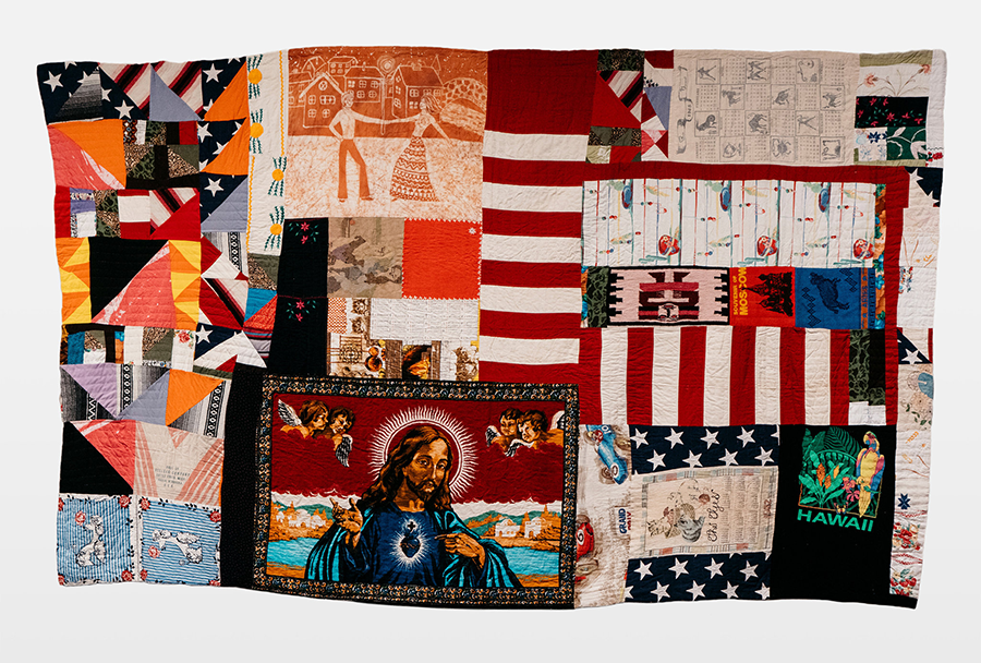 A large quilt comprises a wide array of different squares. Elements of the American flag appear in multiple places; a kitschy image of Jesus appears conspicuously in a bottom square; elsewhere, geometric and floral patterns mix with an overall warm but dramatic color scheme.