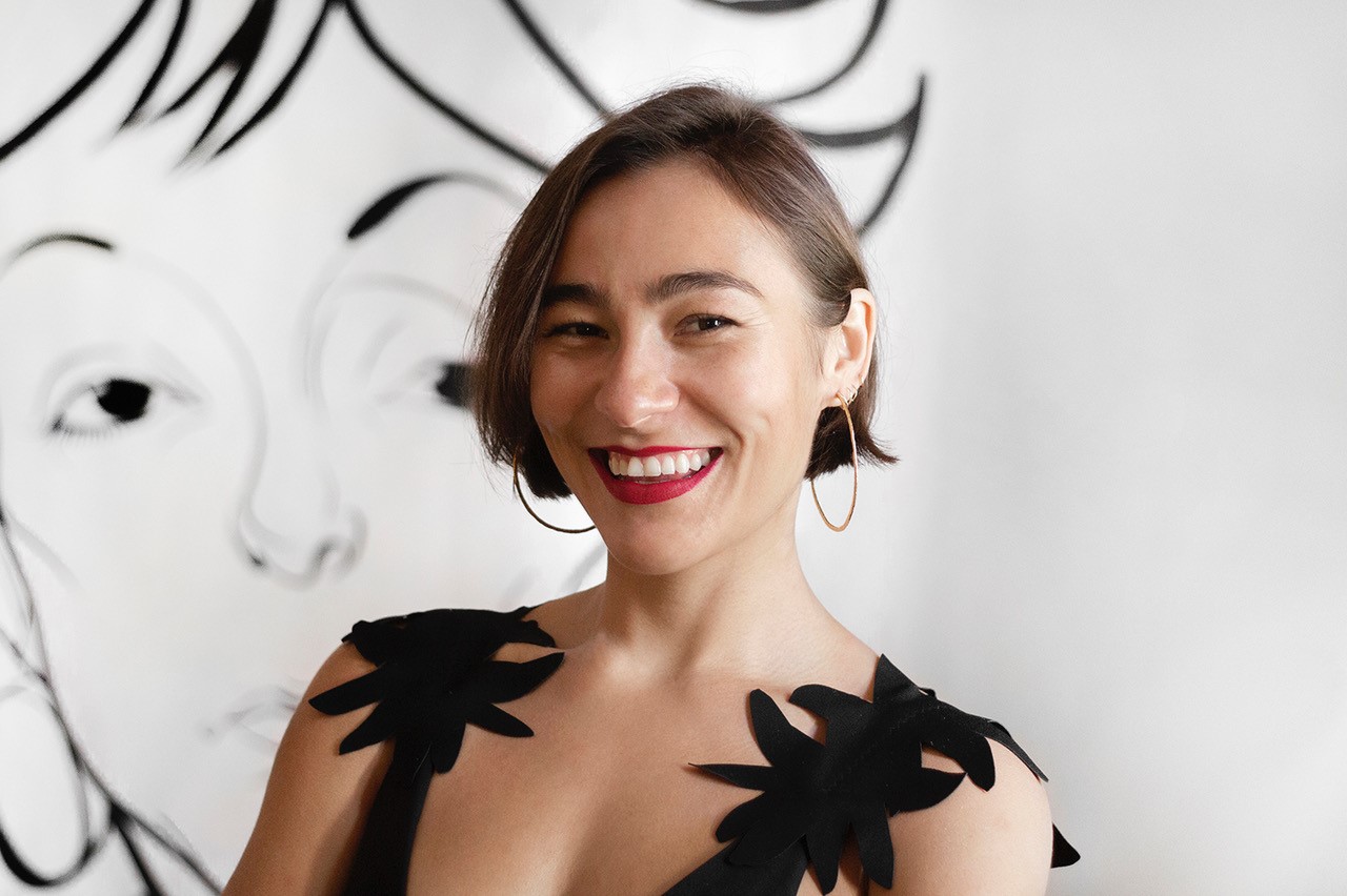 A medium-skinned woman with ear-length straight brown hair smiles widely for the camera. The photograph is cropped from her chest up; she wears a black sleeveless top, red lipstick and gold hoop earrings. Behind her on a white wall the outline of a face, drawn in black outline.