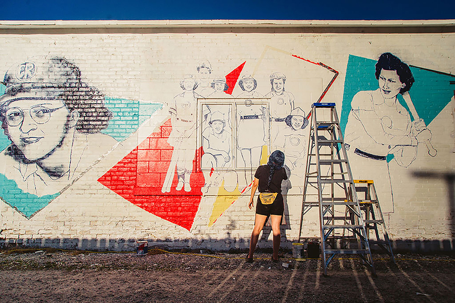 A woman with a yellow fanny pack paints a mural on a white wall in an alley. The mural is unfinished, but shows a group of women in uniforms standing together against a graphic red triangle. Two larger portraits of these women are painted on either side in blue triangles; one is holding a baseball bat. Two ladders stand against the wall.