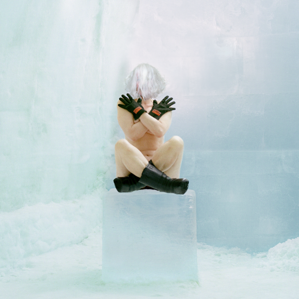 A self-portrait of a light-skinned woman who is naked save for a black hands gloves and black calf-high boots. She sits cross-legged on a block of ice surrounded by walls of ice. Her short white hair hangs in front of her face and her arms are crossed in an "x" over her chest.