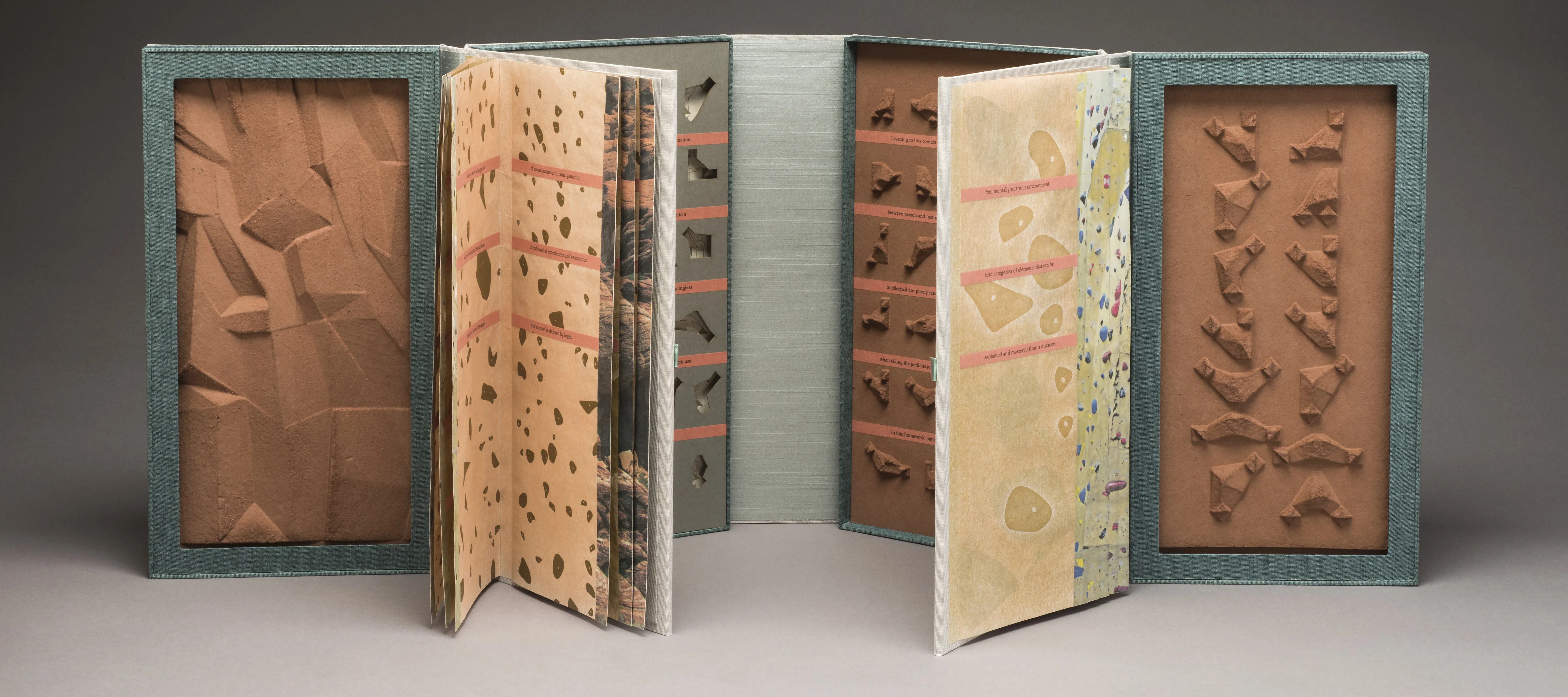 An open artist’s book resembling two open books connected end to end. The four inner cover panels feature small sculpted brown shapes, small indented shapes, and a rocky-looking panel. The two groups of pages feature different patterns that resemble rock climbing walls.