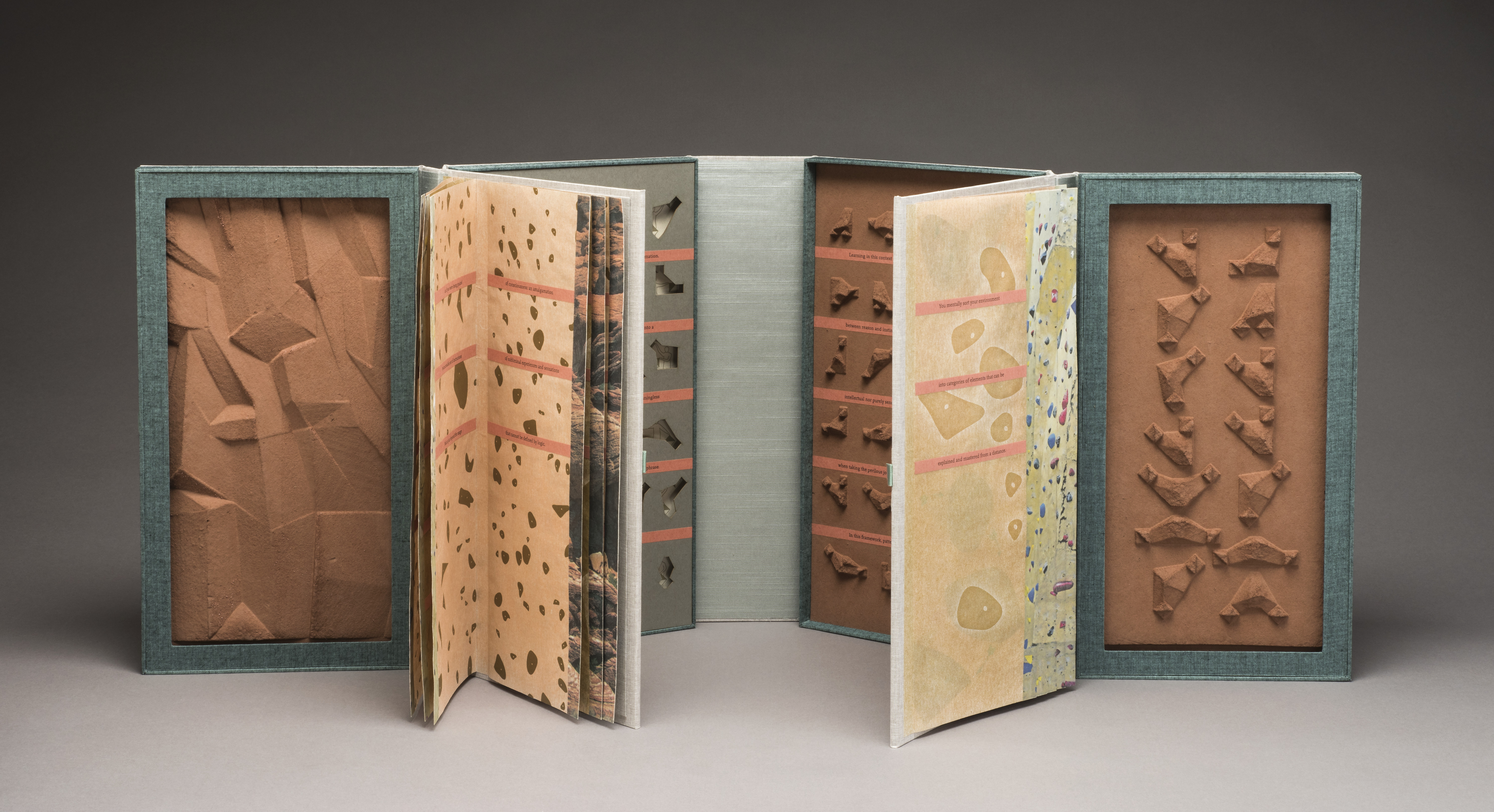 An open artist’s book resembling two open books connected end to end. The four inner cover panels feature small sculpted brown shapes, small indented shapes, and a rocky-looking panel. The two groups of pages feature different patterns that resemble rock climbing walls.