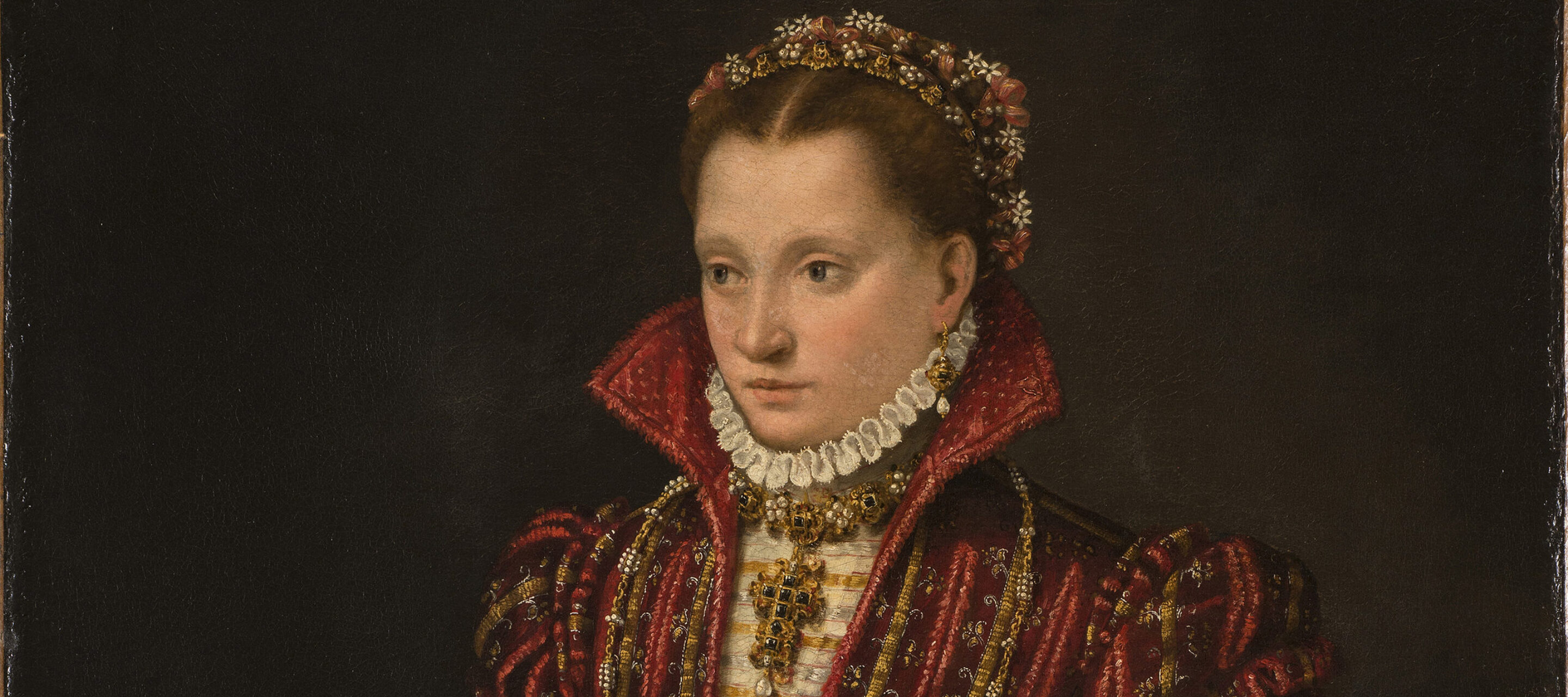 A woman stands in a richly brocaded red dress, her right hand reaching down to pet a small, white dog. Adorned in jewelry, she wears flowers in her hair, parted in the center. The pelt of a small mammal, its head encased in a jeweled holder, hangs from her heavily decorated belt.