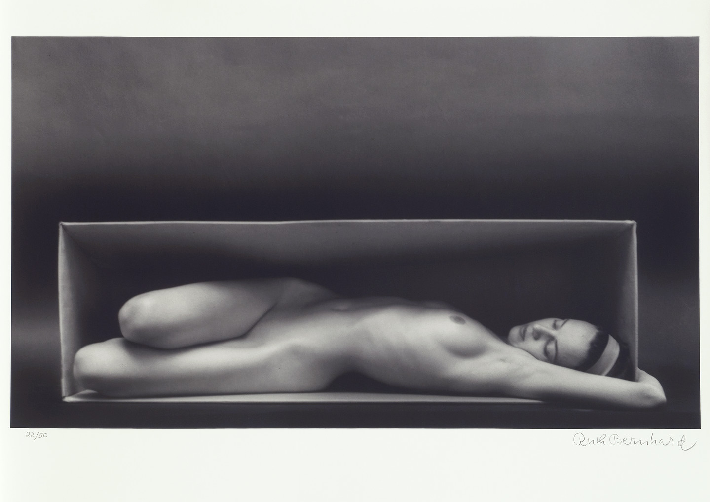 A black-and-white photo of a nude light-skinned woman laying inside the horizontal opening of a box . Her legs are bent at the knees and facing right while the rest of her body is reclined, her left breast exposed to the camera and left extended arm overhead, wrapped around the side of the box. She looks relaxed, with closed eyes and wears a headband. 