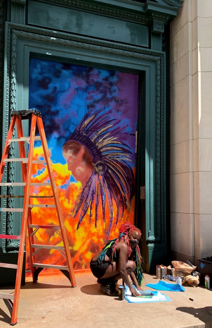 A dark-skinned woman gathers materials on the ground in front of a doorway. She has long bright orange and red hair accentuated with blue and green feathers. In the doorway is a vibrant mural with the profile of a dark-skinned person wearing a sweeping feathered headdress.