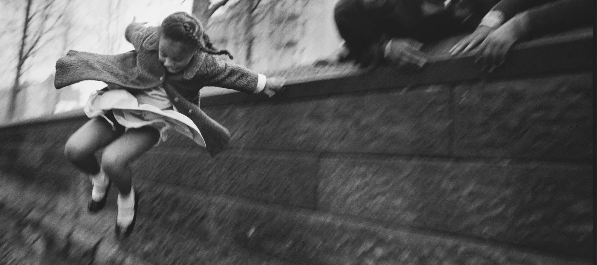 A black and white photograph of a young girl wearing a dress and coat jumping over a brick wall. Three other figures lean over the wall in the upper right corner of the composition. The jumping girl is the only figure who is in focus.