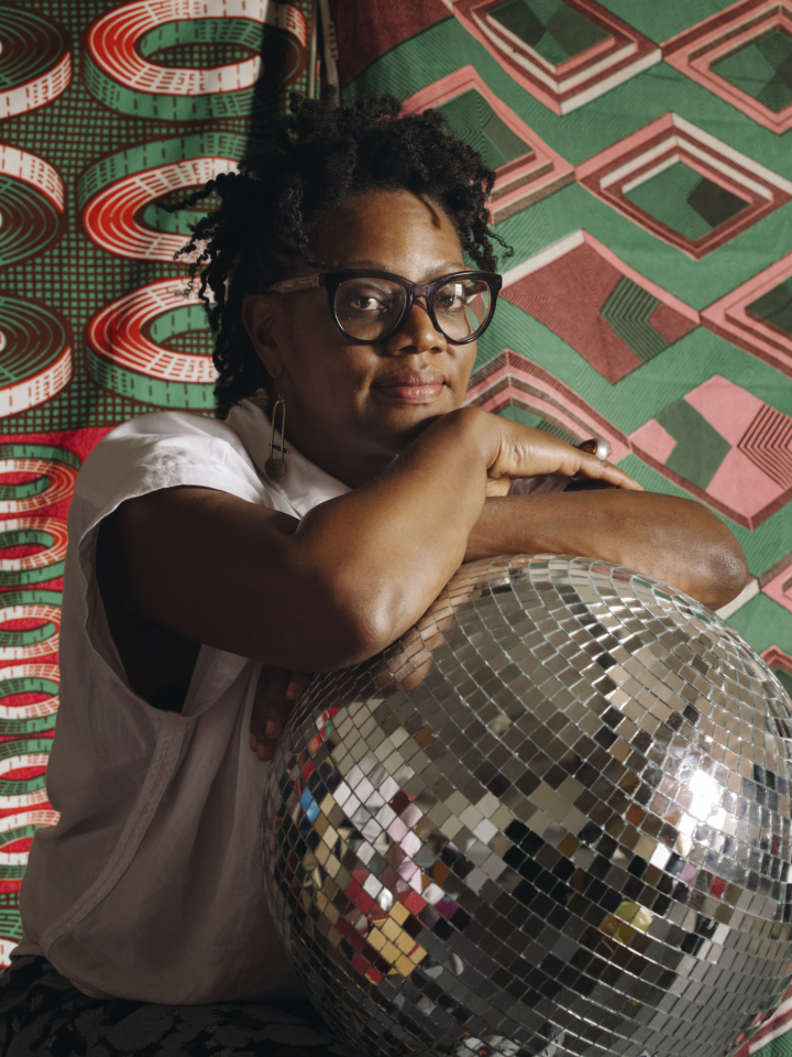 An African American woman sits in front of a green and red lively-patterned background; she stares slightly smiling at the camera, her arms resting on the top of a large silver disco ball and just below her chin. She wears large cat-eye glasses and a white shirt. 