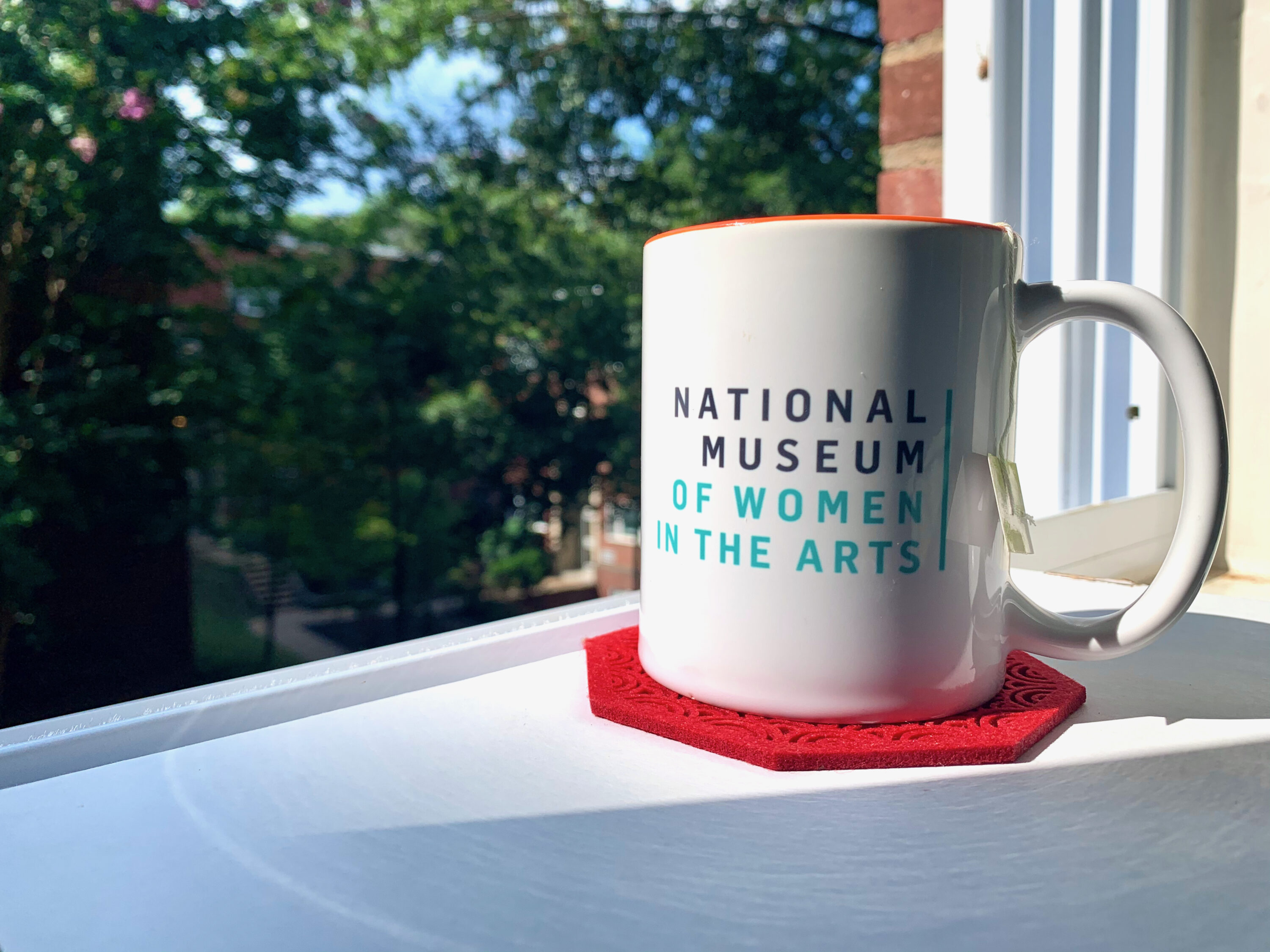 A white mug with National Museum of Women in the Arts printed on it in gray and green text, sitting on a red coaster on a white surface. Behind the mug is an open window showcasing green foliage.