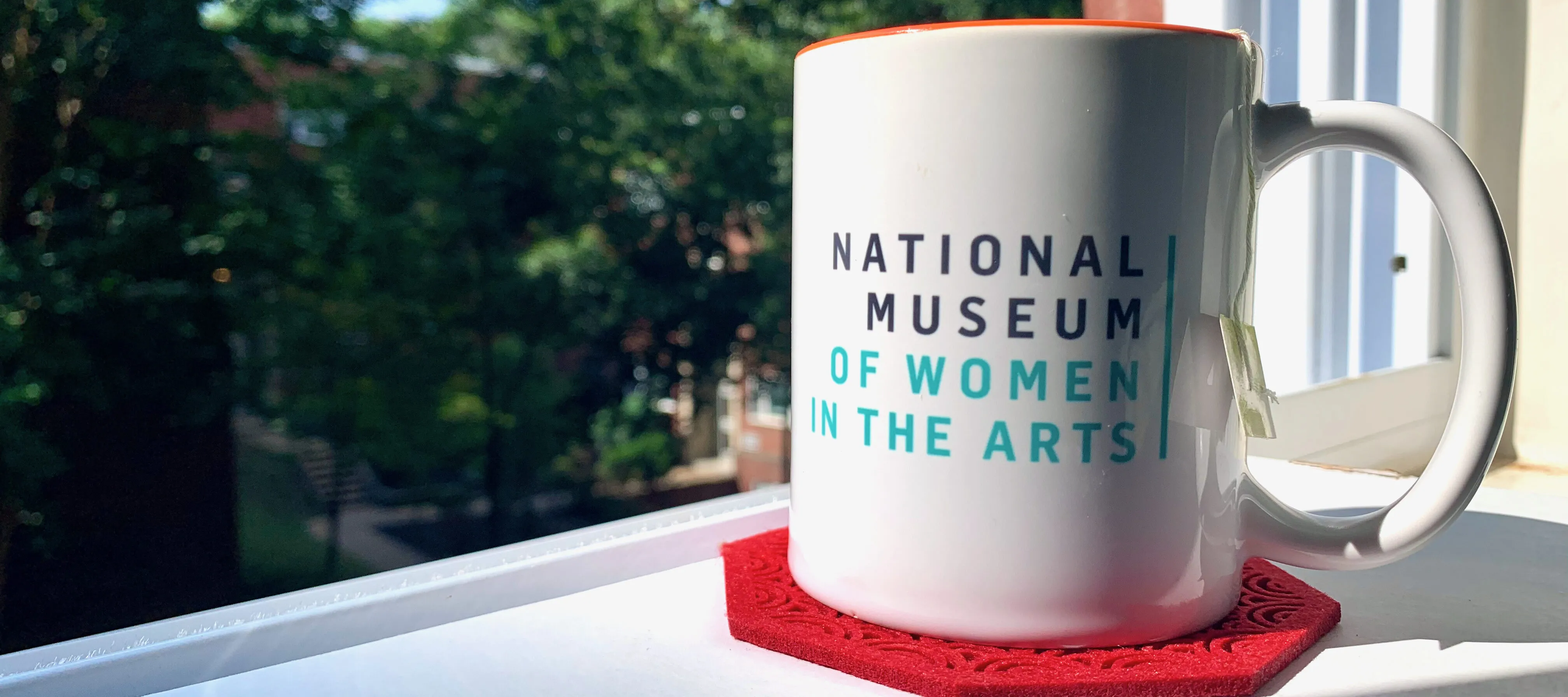 A white mug with National Museum of Women in the Arts printed on it in gray and green text, sitting on a red coaster on a white surface. Behind the mug is an open window showcasing green foliage.