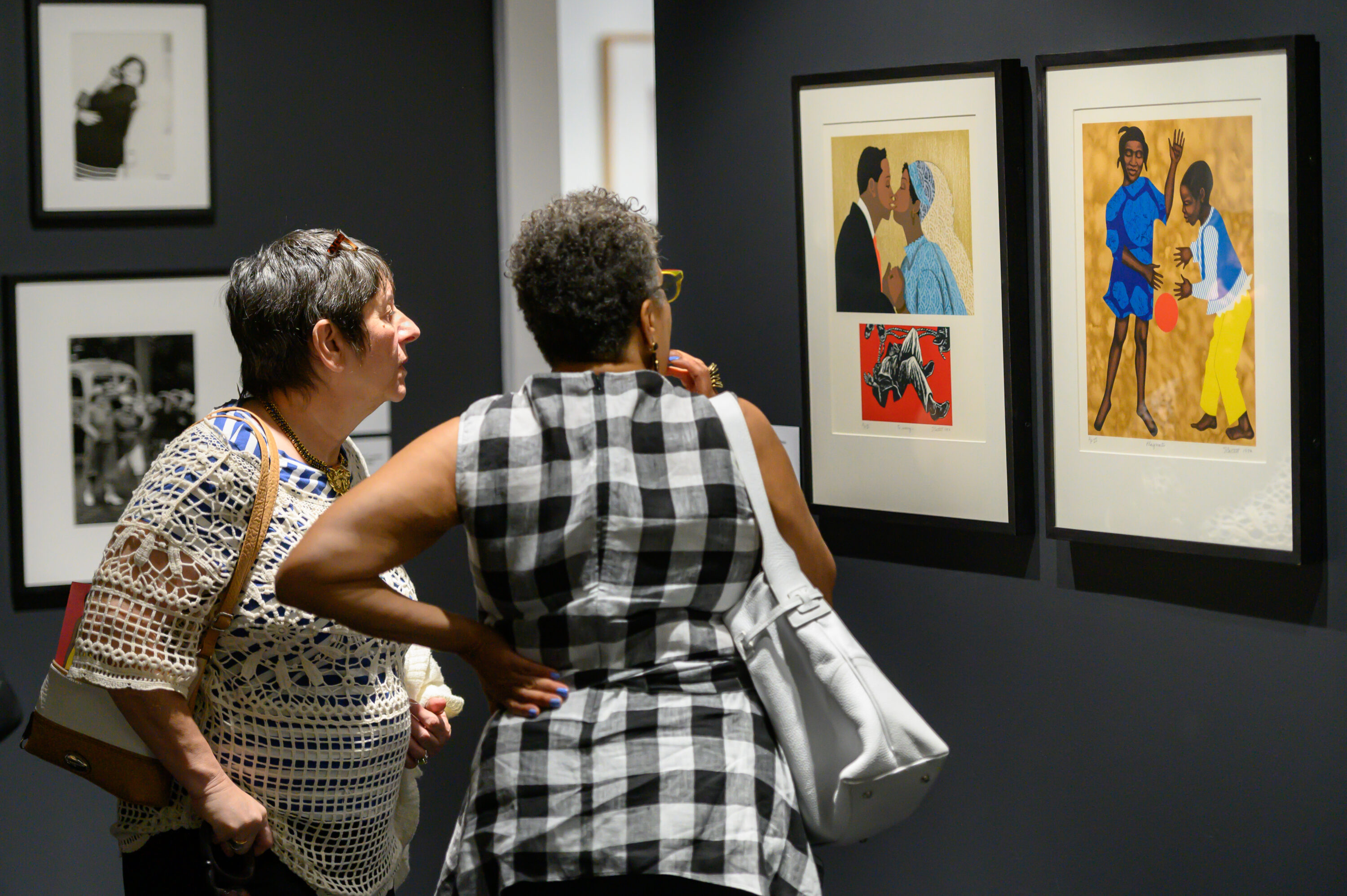 A light-skinned older woman with a cropped hair cut and a dark-skinned older woman also with a cropped hair cut stand in a gallery looking at two colorful lithographs framed side-by-side on a dark grey wall. The artwork on the left shows a dark-skinned couple mid-kiss after getting married; the artwork on the right shows a dark-skinned girl and boy playing with an orange ball. 