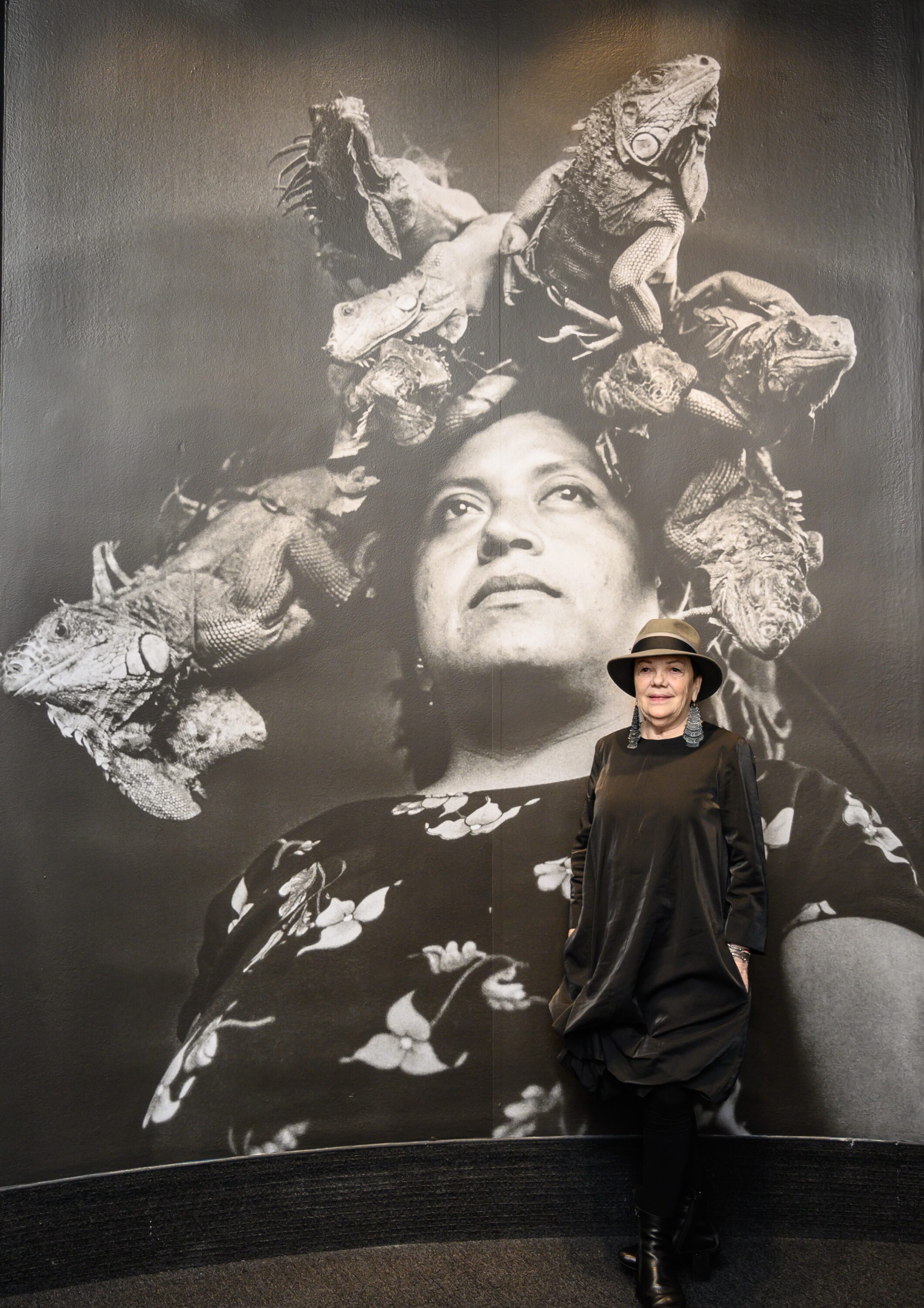 A light-skinned woman wearing a brimmed hat, geometric earrings, and a long-sleeved black velvet dress stands in front of a wall-sized black and white portrait of a woman, who has eight iguanas perched on her head like a crown. The photographed woman looks content and confident.