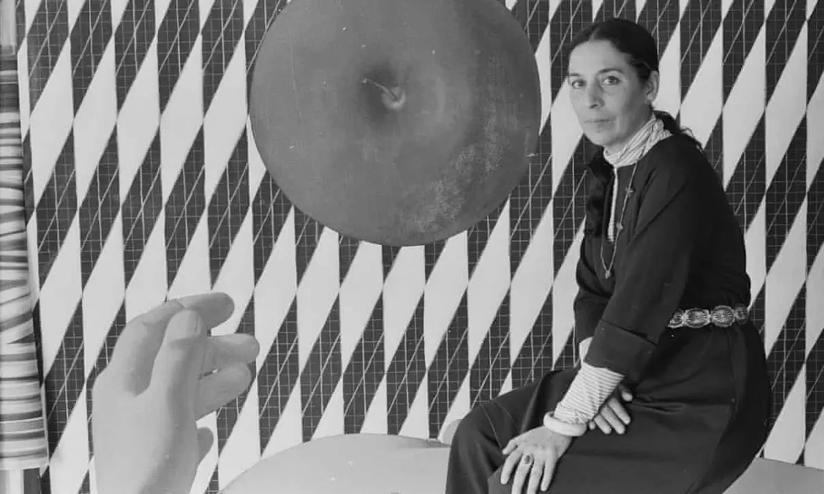 A black-and-white photo of a light-skinned woman sitting in front of a geometric abstract painting that features vertical diamond shaped with a grid-like pattern. A large top-view of an apple is the central focus and a human hand emerges from the bottom of the frame.