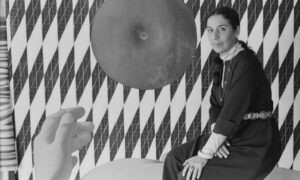 A black-and-white photo of a light-skinned woman sitting in front of a geometric abstract painting that features vertical diamond shaped with a grid-like pattern. A large top-view of an apple is the central focus and a human hand emerges from the bottom of the frame.