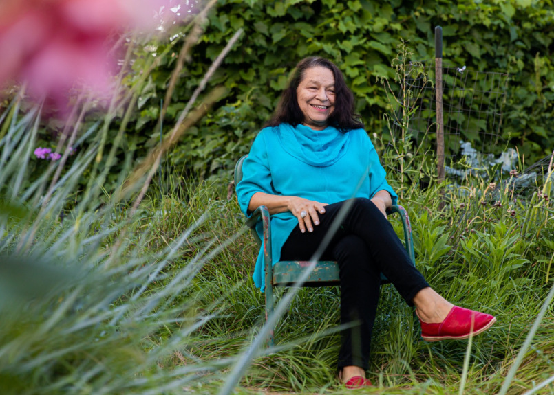 A Native American woman sits in a rusted metal chair in a wooded, grassy setting. She wears a loose-fitting bright blue shirt, black pants, and red loafers; she smiles brightly and looks to the left of the camera, her hands resting casually on her legs.