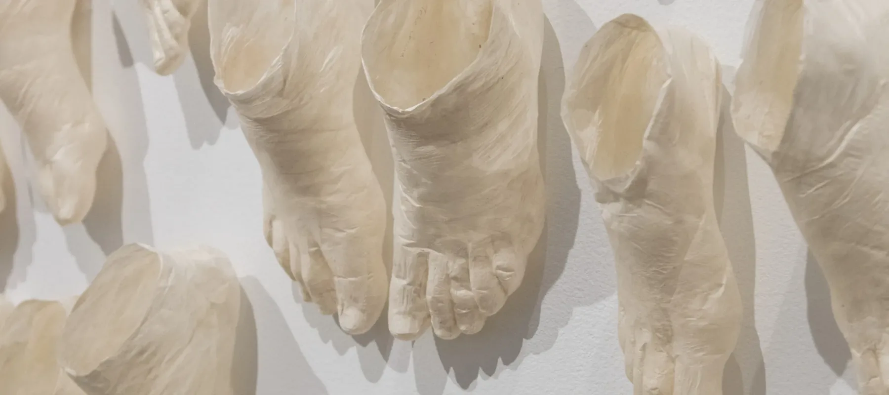 Detail view of a contemporary art installation made up of countless beige foot sculptures that extend from the walls onto the dark floor. The feet are in pairs in varied sizes and staggered as if moving. Marks on the sculptures indicate the folds of human skin and bone.