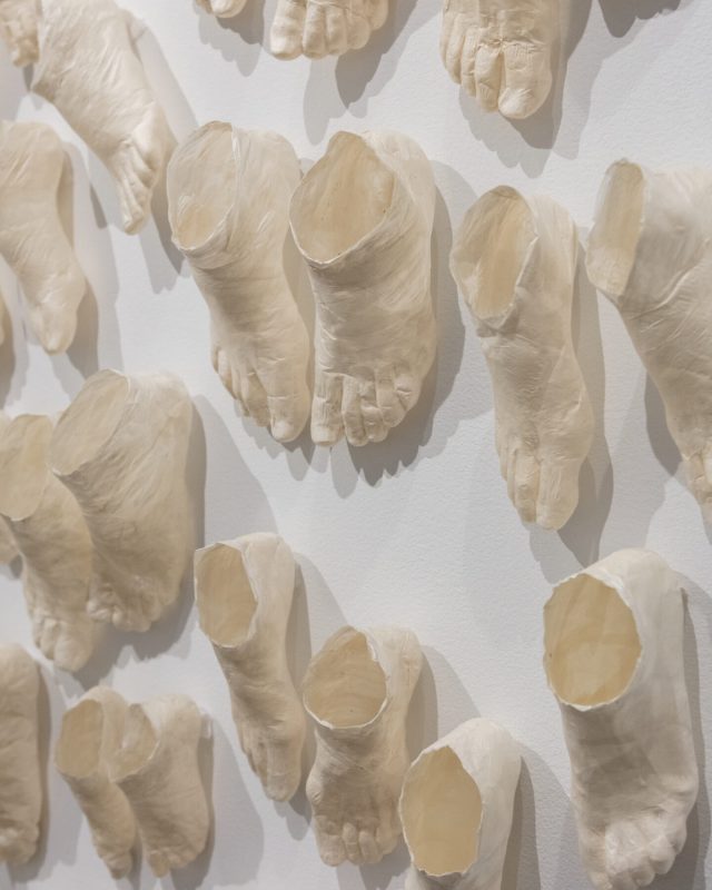 Detail view of a contemporary art installation made up of countless beige foot sculptures that extend from the walls onto the dark floor. The feet are in pairs in varied sizes and staggered as if moving. Marks on the sculptures indicate the folds of human skin and bone.