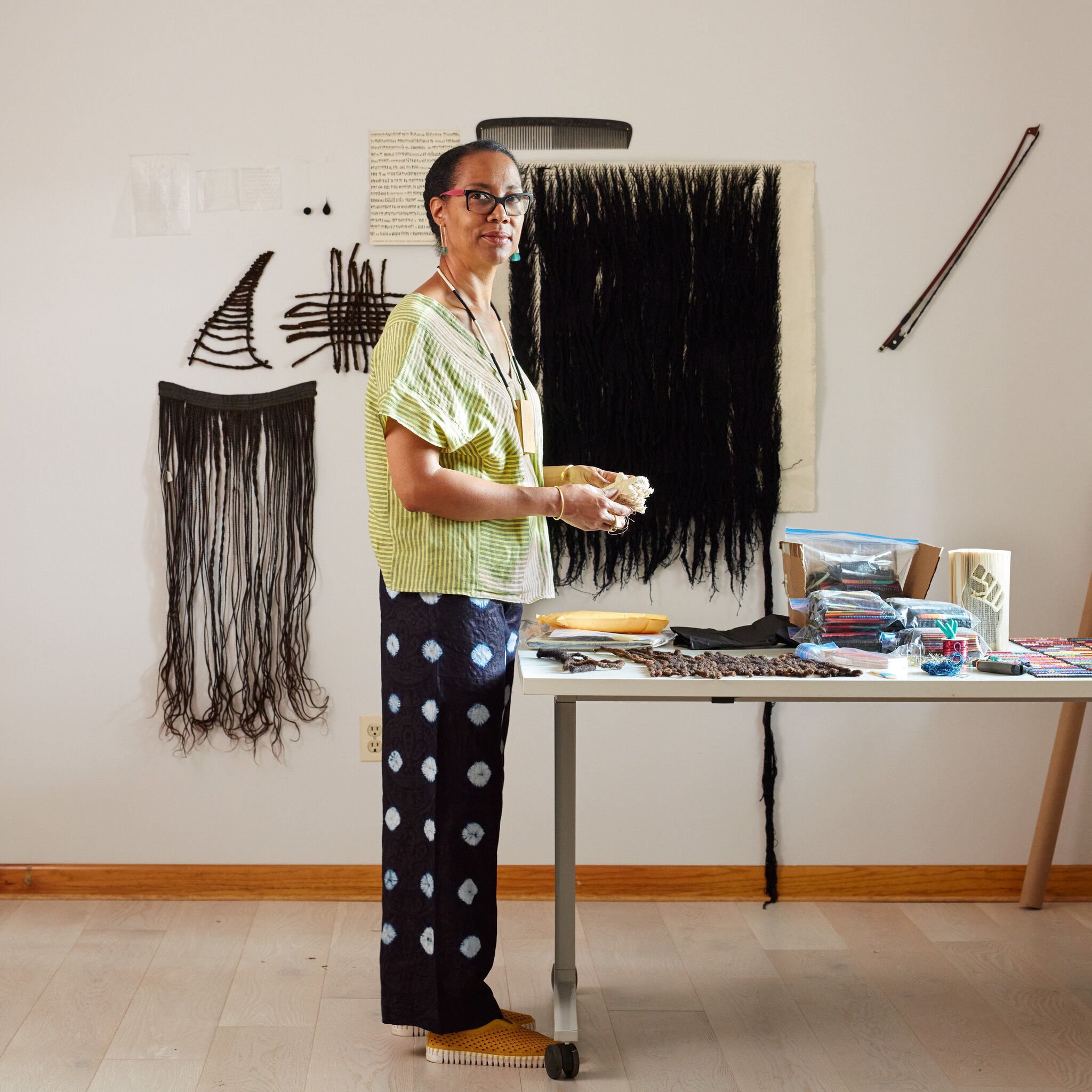 A dark-skinned woman stands facing the right next to a worktable in an art studio. She looks directly at the camera; her hands hold a piece of material in front of her. On the wall behind her is a over-sized comb hung above four distinctly different tapestries comprised of strands of black hair.
