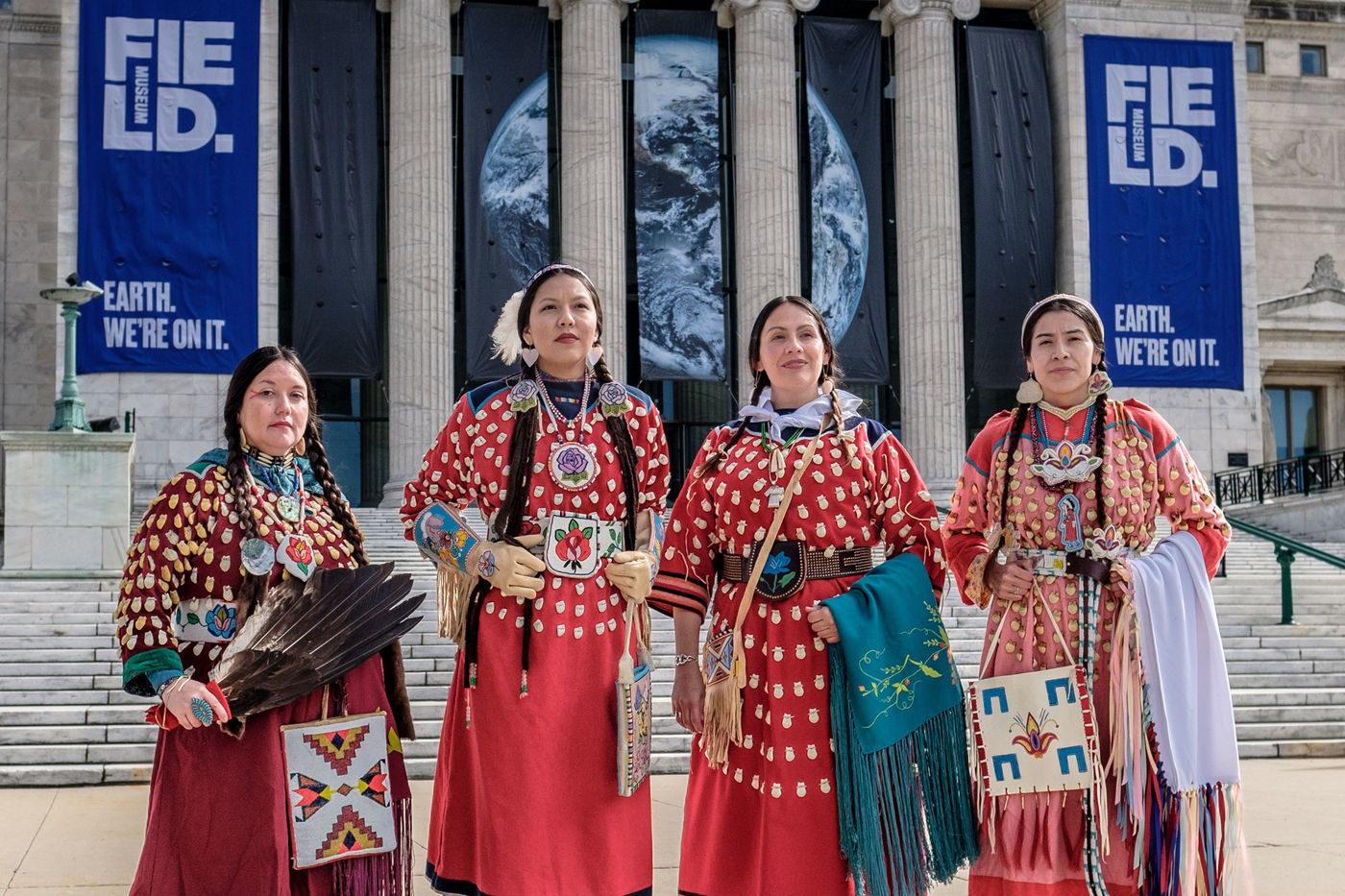 Four women stand with confidence in front of a grand marble museum façade. They each wear a long red dress with gold dot patterns and hold various bags, blankets, and belts with distinctive geometric patterns. Each woman’s long brown hair is braided in two low pigtails.