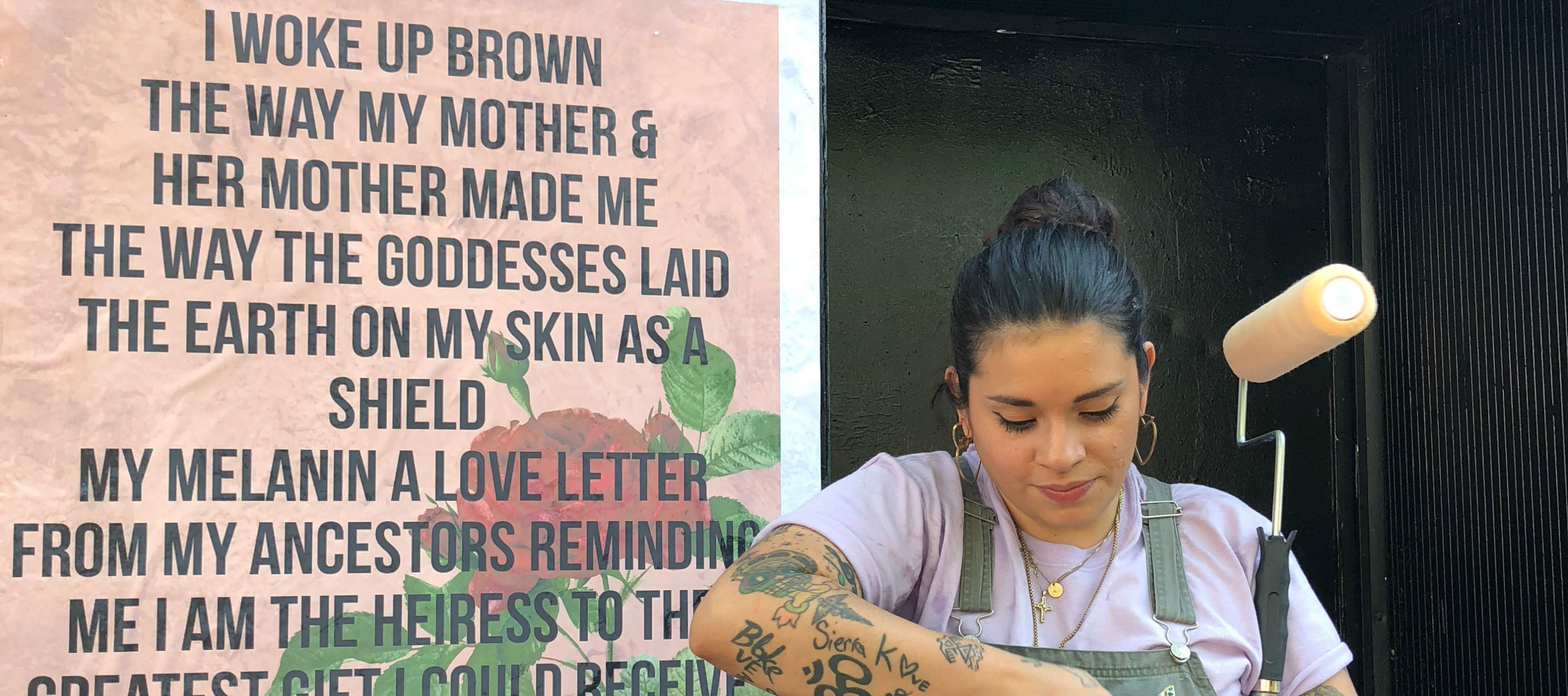 A woman holds a paint roller and looks down, her hands at work; She stands next to a wheat paste mural that features bold black text overlaid on a peach background that features a rose. The text reads: 