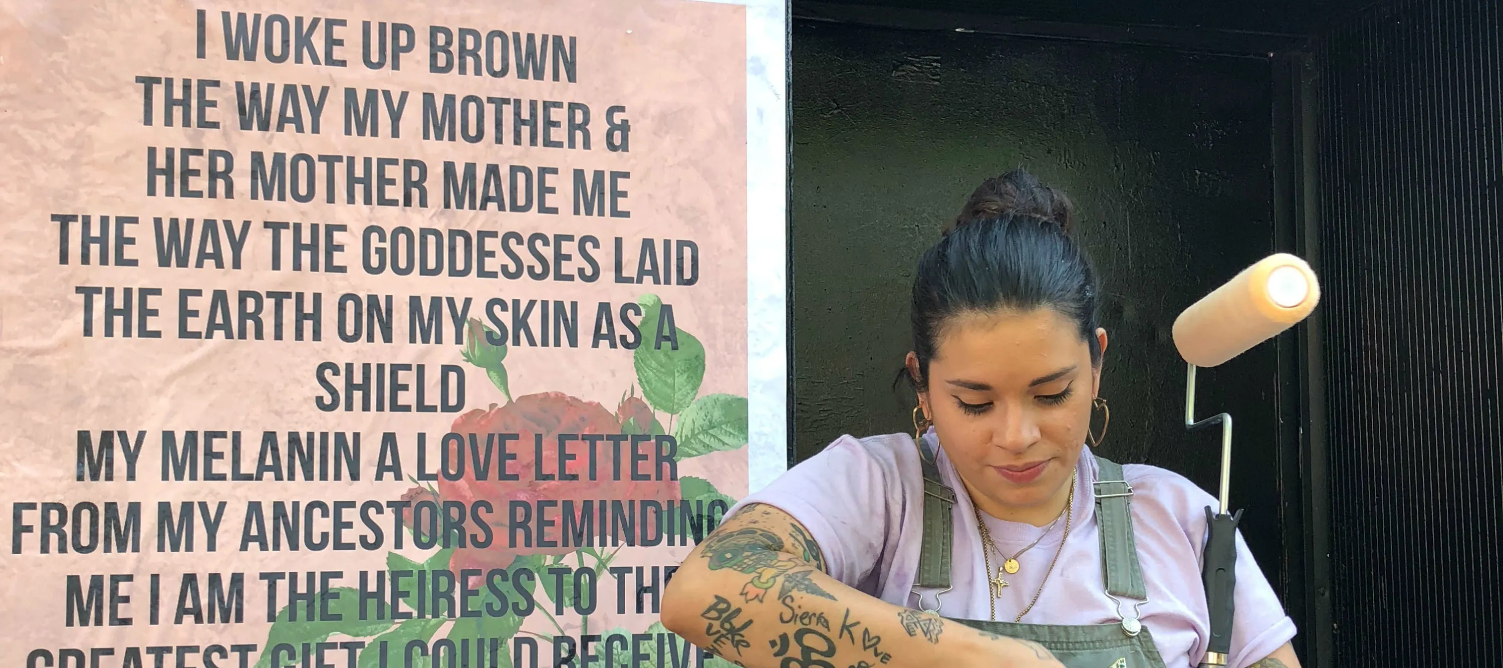 A woman holds a paint roller and looks down, her hands at work; She stands next to a wheat paste mural that features bold black text overlaid on a peach background that features a rose. The text reads: "I woke up Brown/The way my mother &/Her Mother made me/The way the goddesses laid/The earth on my skin as a shield/My melanin is a love letter/From my ancestors reminding/Me I am the heiress to the/Greatest gift I could receive/The crown of the sun.