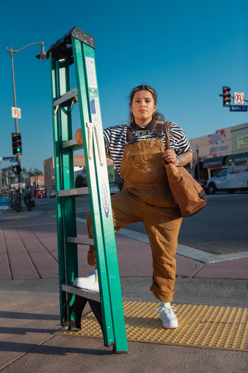 A woman wearing camel colored overalls stands on a city street corner, one arm holding a shoulder bag and the other rm and leg holding a tall green ladder vertical.