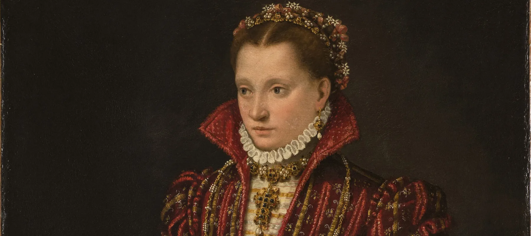 A woman stands in a richly brocaded red dress, her right hand reaching down to pet a small, white dog. Adorned in jewelry, she wears flowers in her hair, parted in the center. The pelt of a small mammal, its head encased in a jeweled holder, hangs from her heavily decorated belt.