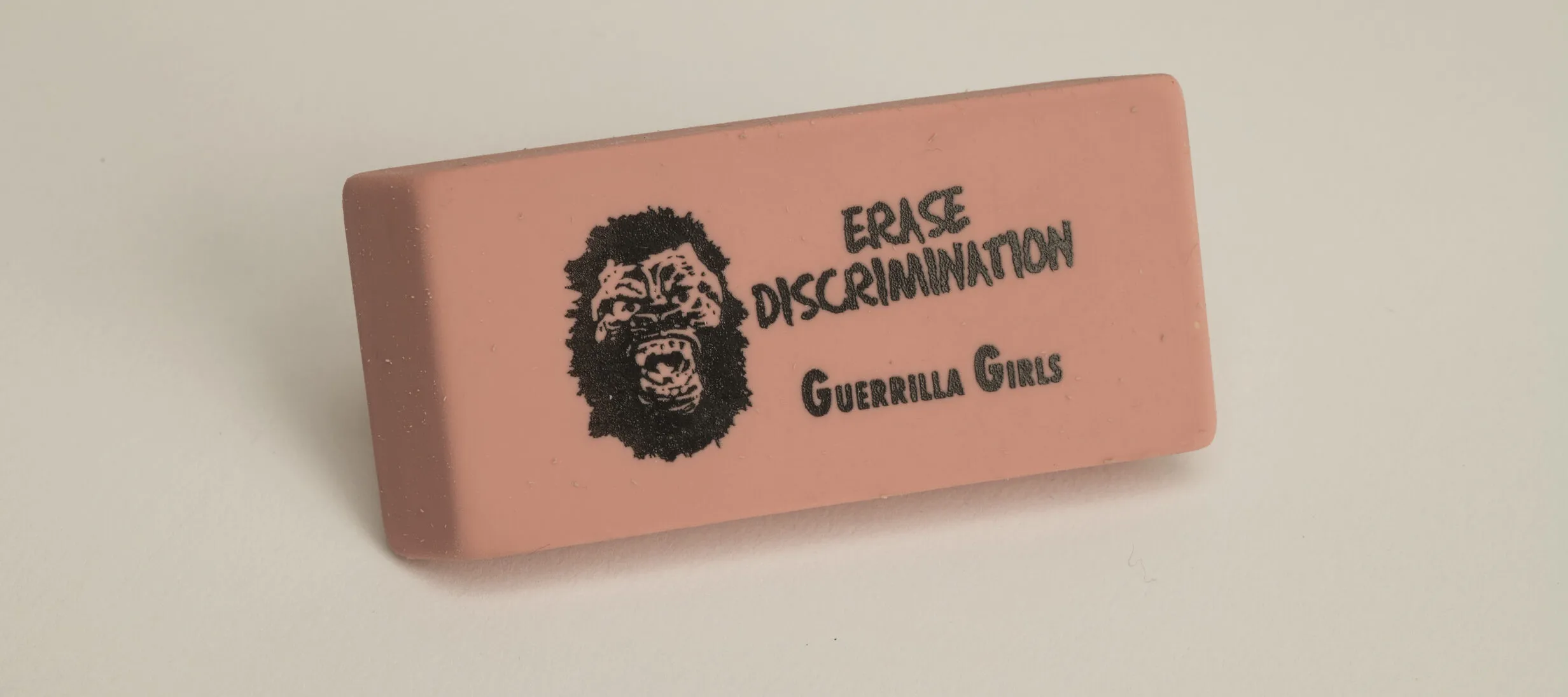 A rectangular pink eraser against a white background. Printed on the eraser in black is a yelling gorilla face to the left of the words, "ERASE DISCRIMINATION" above the words "Guerrilla Girls."