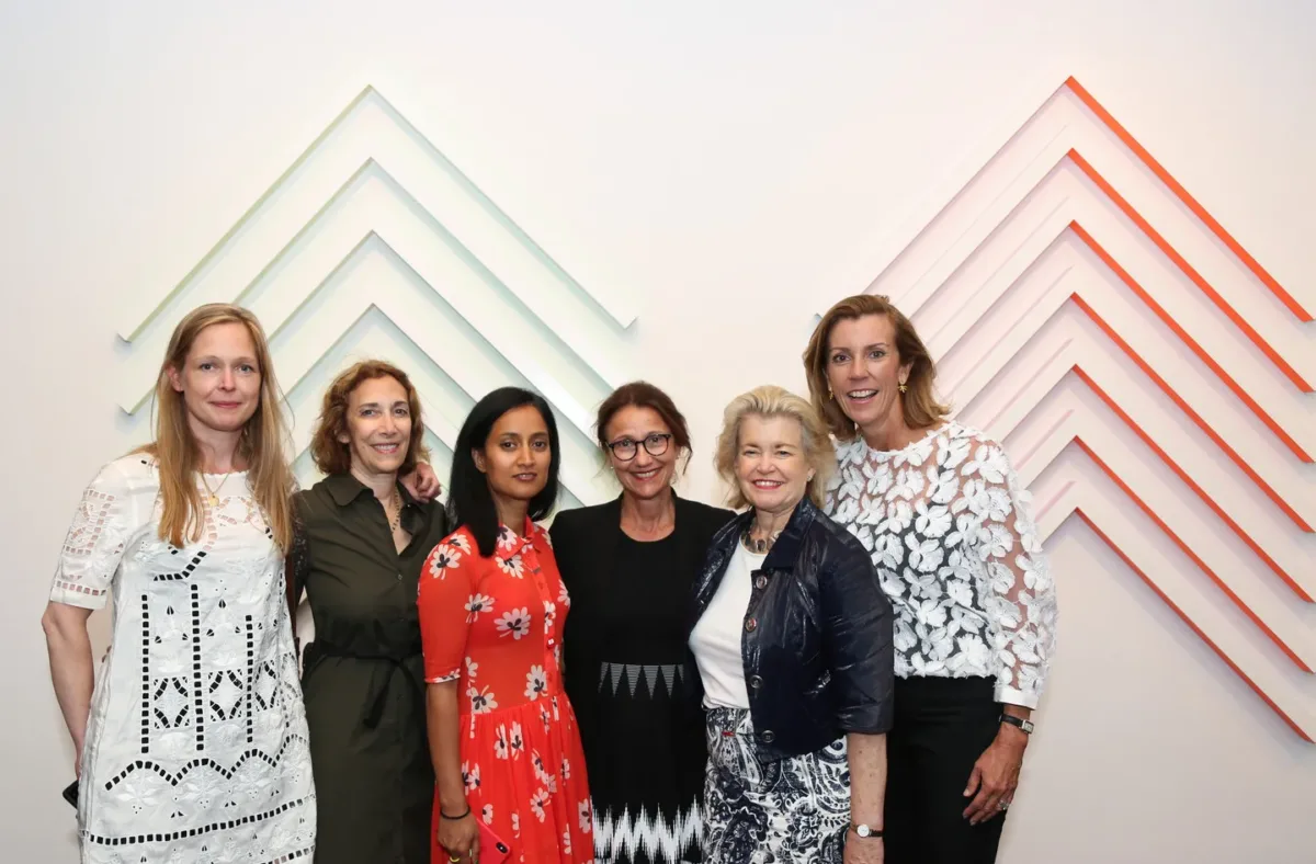 A photograph of a group of five light-skinned women and one medium-skinned woman dressed in cocktail attire stand in front of a geometric wall sculpture.