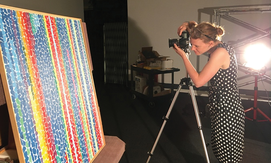 A light-skinned woman with blonde hair wearing a sleeveless polka-dot dress stands in a dark gallery behind a camera and tripod. She photographs an abstract painting composed of brightly colored, lozenge-shaped brushstrokes in vertical stripes of rich blue, purple, turquoise, yellow, orange and red.