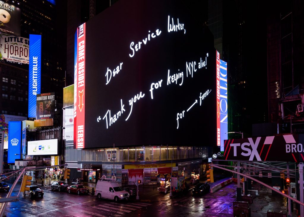 In Times Square, New York City, a large digital billboard features a black background and white handwritten text that says "Dear Service Worker, Thank you for keeping NYC alive!" for ---> forever...