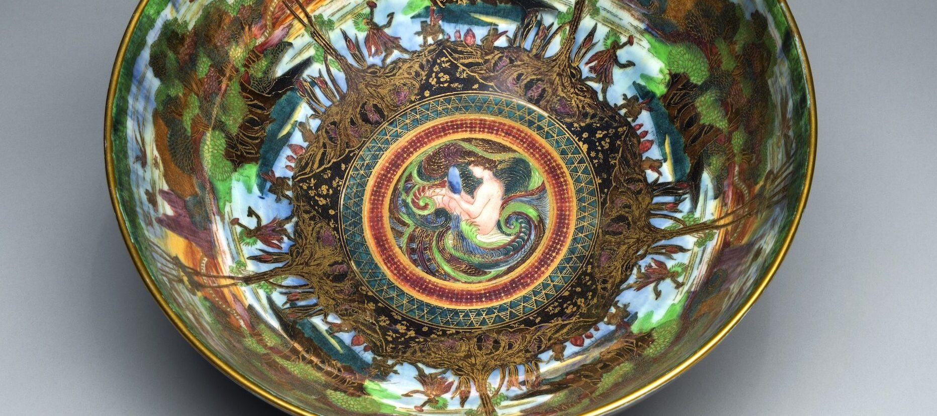 Interior view of a colorful, iridescent lusterware bowl featuring gold cloisonne and an ornamental Art Nouveau design. At the center, a light-skinned mermaid bathes in the ocean. Radiating outward, and repeated five times, is a lush agrarian scene.
