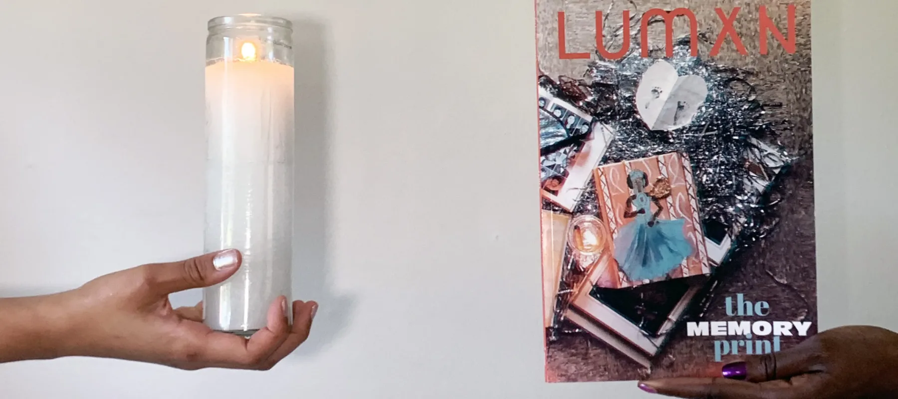 Two hands enter the frame from the left and right sides. On the left, the light-skinned hand has silver nail polish and holds a white, tall, lit votive candle. The hand on the right is dark-skinned and holds a magazine that says "LUMXN: The Memory Print" and features a collage on the cover.