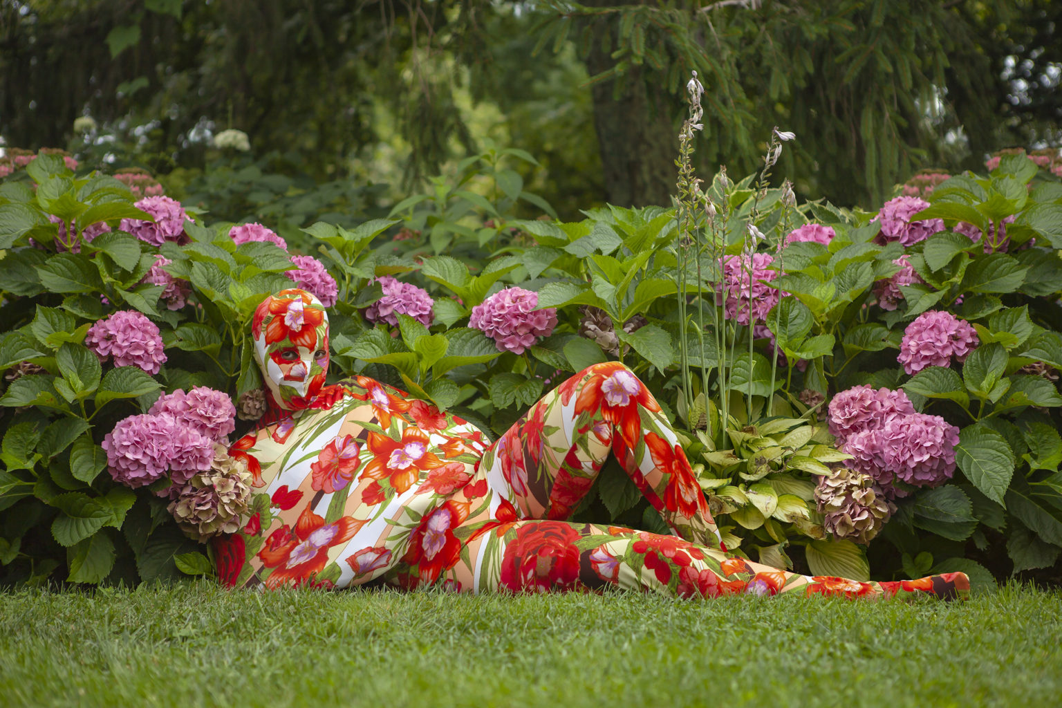 A figure wearing a tight body suit and head mask covered in a red and pink floral print reclines in cut grass, in front of blooms of purple flowers. Only the eyes of the figure are visible.