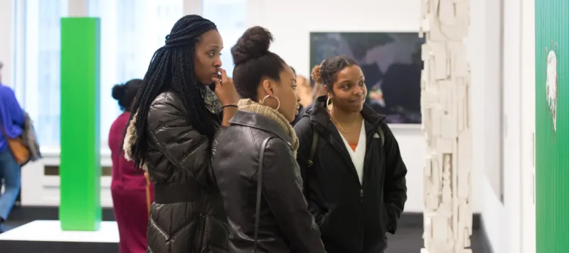 Three dark-skinned young adults in winter jackets stand in an art gallery near a white pillar made of a medley of found items. They wear black winter jackets and smile at the artwork.