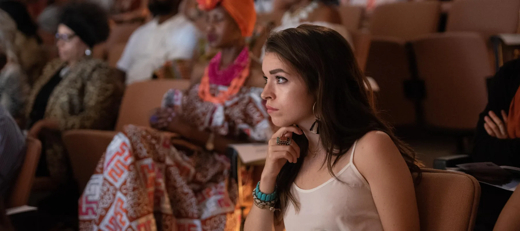 An audience seated in a dim theater looking intently forward. Focused in the foreground is a light-skinned person with their chin on their hand, long brown hair, and a tank top.