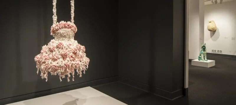 A sculpture hangs in a dark gallery. The sculpture comprises layers of melted pink and white wax that form a dress-like shape hanging from satin-wrapped chains. Its color, shape, and bumpy, lacy texture, evoke a frilly tutu, lavishly frosted wedding cake, or coral.