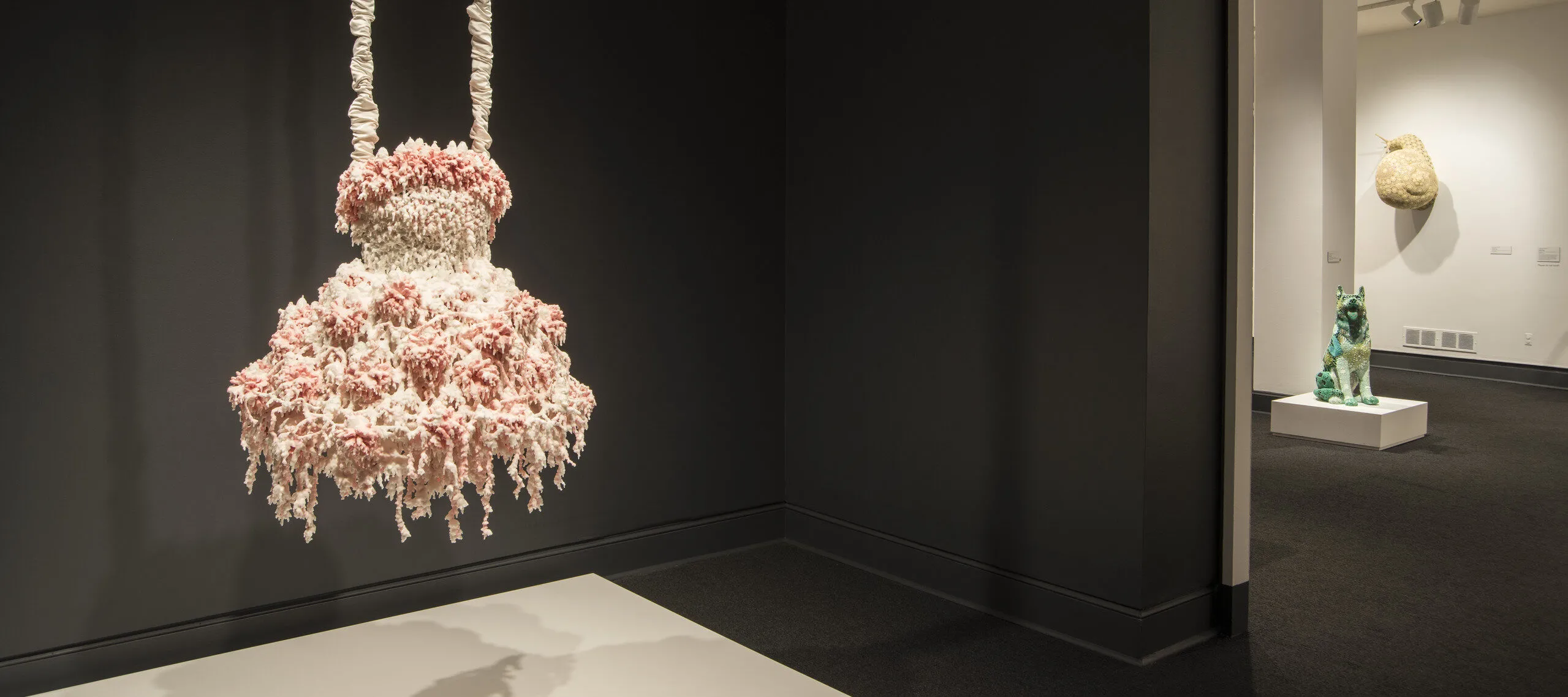 A sculpture hangs in a dark gallery. The sculpture comprises layers of melted pink and white wax that form a dress-like shape hanging from satin-wrapped chains. Its color, shape, and bumpy, lacy texture, evoke a frilly tutu, lavishly frosted wedding cake, or coral.