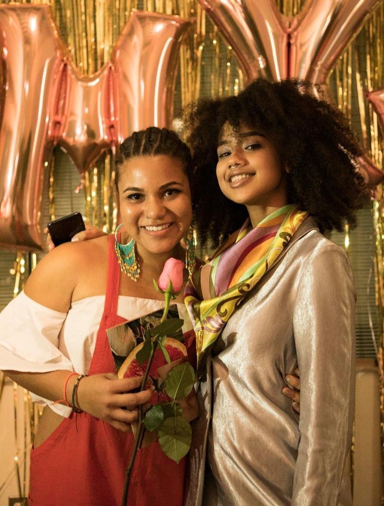 Two dark-skinned women lean close together and smile at the camera in front of shiny rose gold balloons and gold fringe streamers. The woman on the left holds a pink rose and a book that features a photo of a grapefruit half. She has her arm around the waist of the woman on the right, who wears a velvety blazer and pastel patterned scarf.