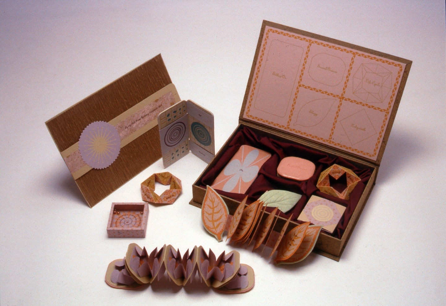Multiple colorful objects sit spread out against a neutral background, including a rectangular box filled with more small objects, two small books spread open, a small box resembling a game, and other folded paper objects.