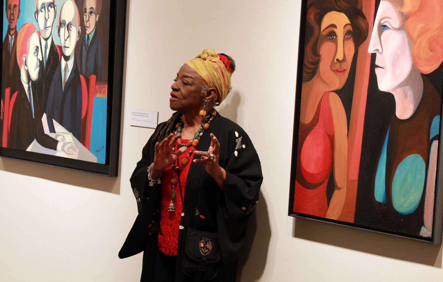 Faith Ringgold stands and speaks, gesturing with her hands, in front of her portrait paintings. She is a dark-skinned adult woman wearing colorful clothing and a black sweater, including a yellow head wrap and chunky jewelry—rings, earrings, necklaces, and bracelets.