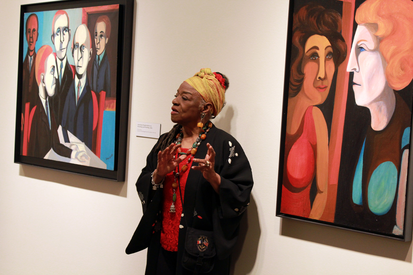 Faith Ringgold stands and speaks, gesturing with her hands, in front of her portrait paintings. She is a dark-skinned adult woman wearing colorful clothing and a black sweater, including a yellow head wrap and chunky jewelry—rings, earrings, necklaces, and bracelets.