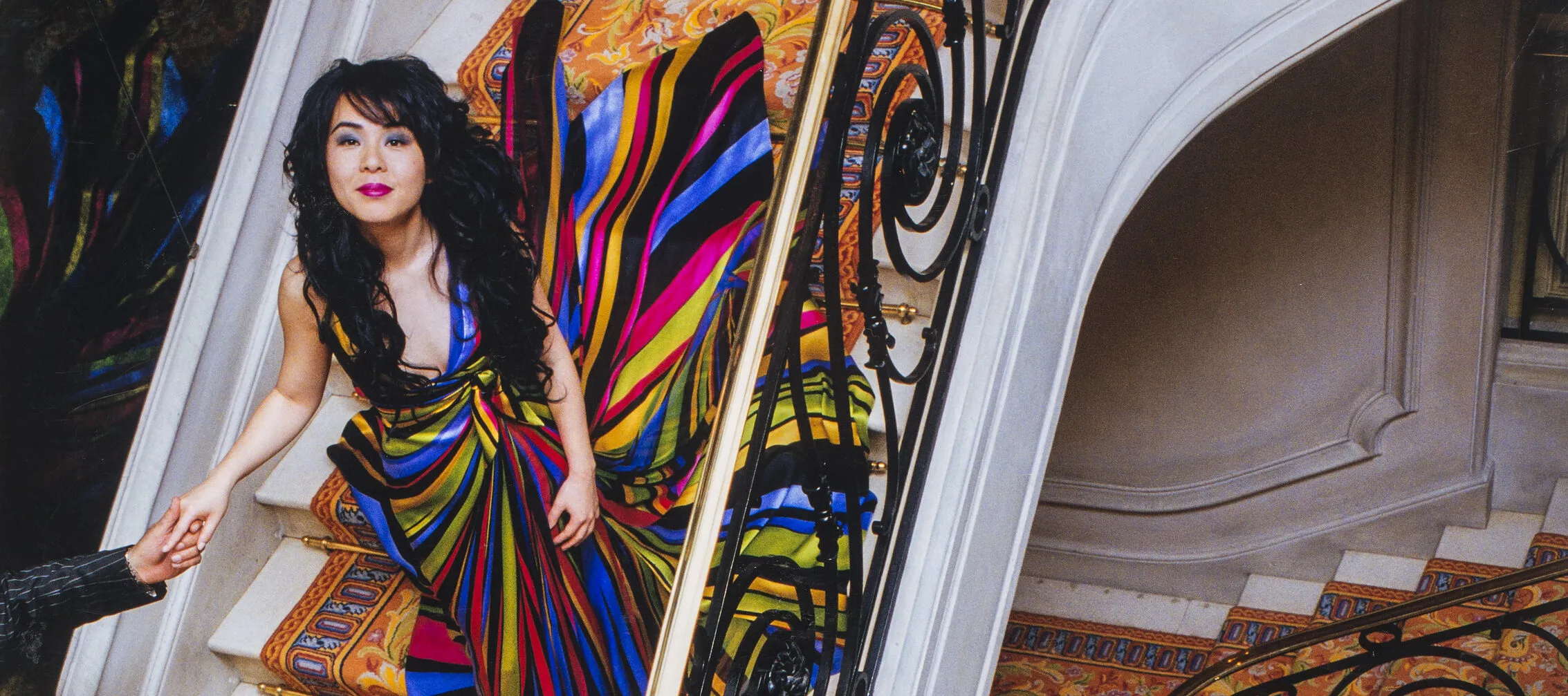 A bird's eye view of a woman with long, dark hair in a brightly-colored gown descending an ornate spiral staircase of white marble and colorfully patterned rug. Her right hand is being held by a figure in a black suit whose head and shoulders are cropped out of the image.