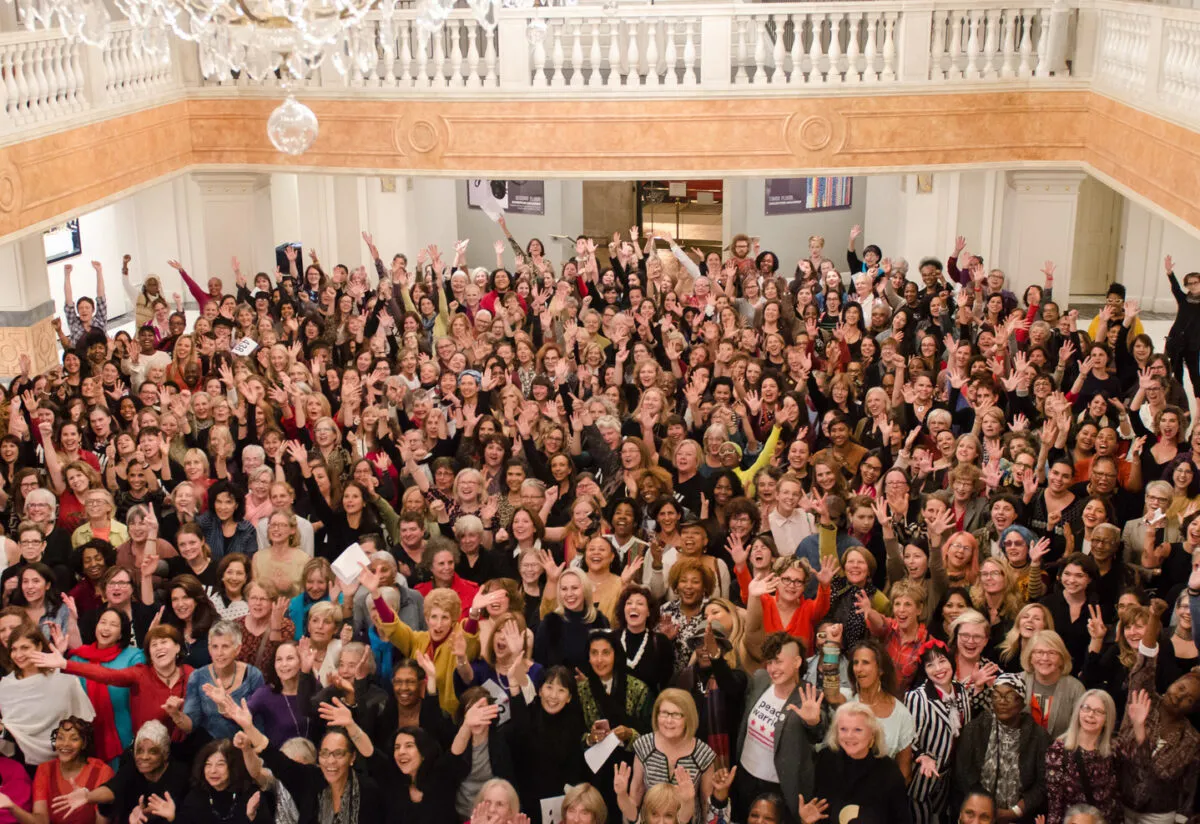 A color photograph of 435 women from a high vantage point. The women all look up towards the camera, some with arms outstretched.