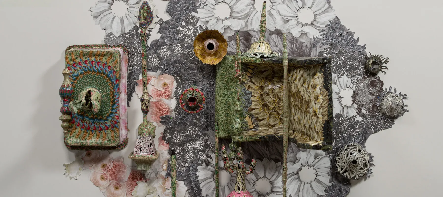 A contemporary wall sculpture with cut wallpaper piece pasted onto the wall and objects and home furniture protrude from the wall and onto the ground covered in colorful floral wallpaper.