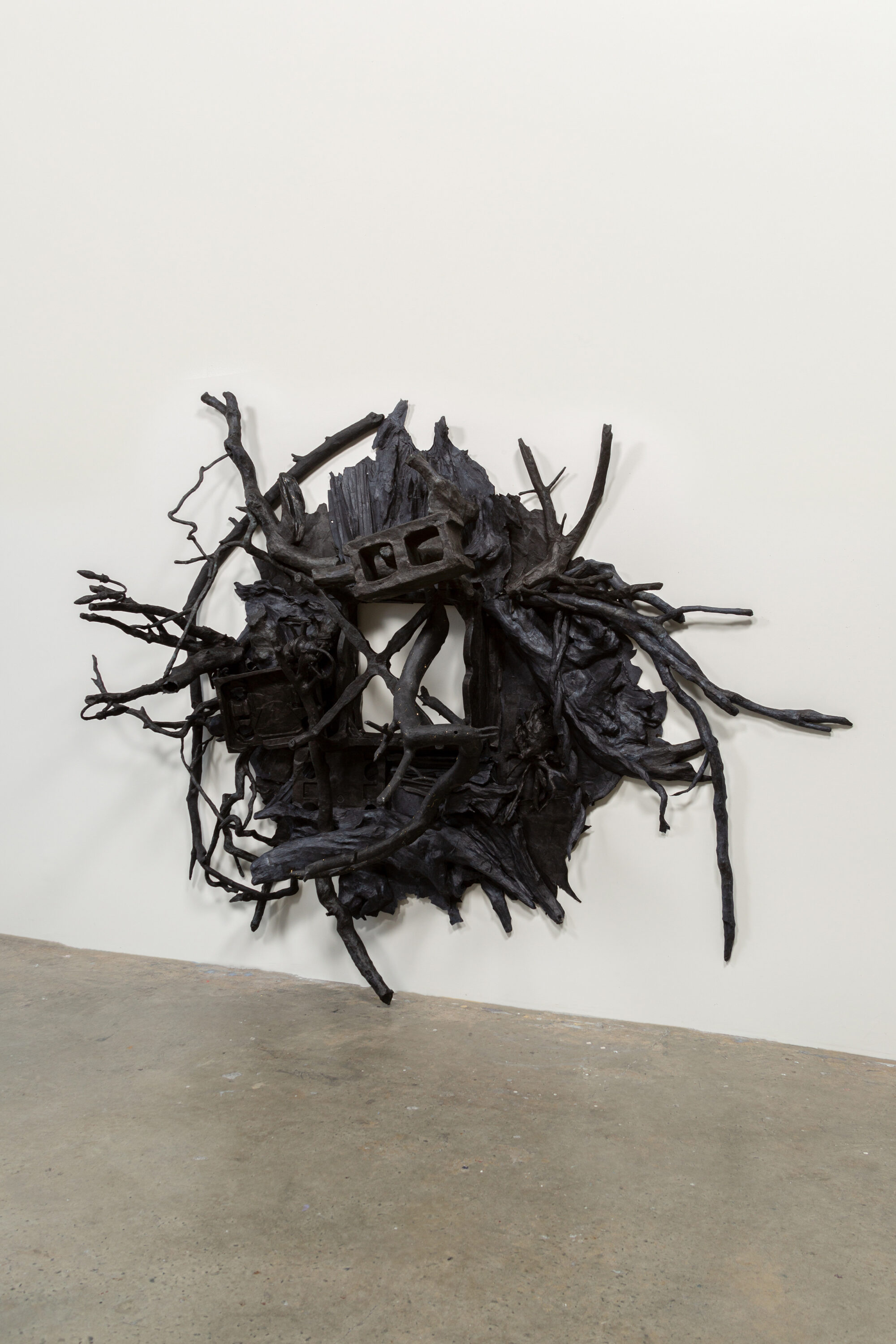 A contemporary black wall sculpture in a rounded shape with many twisted branches protruding out from the work. A void space is in the center.